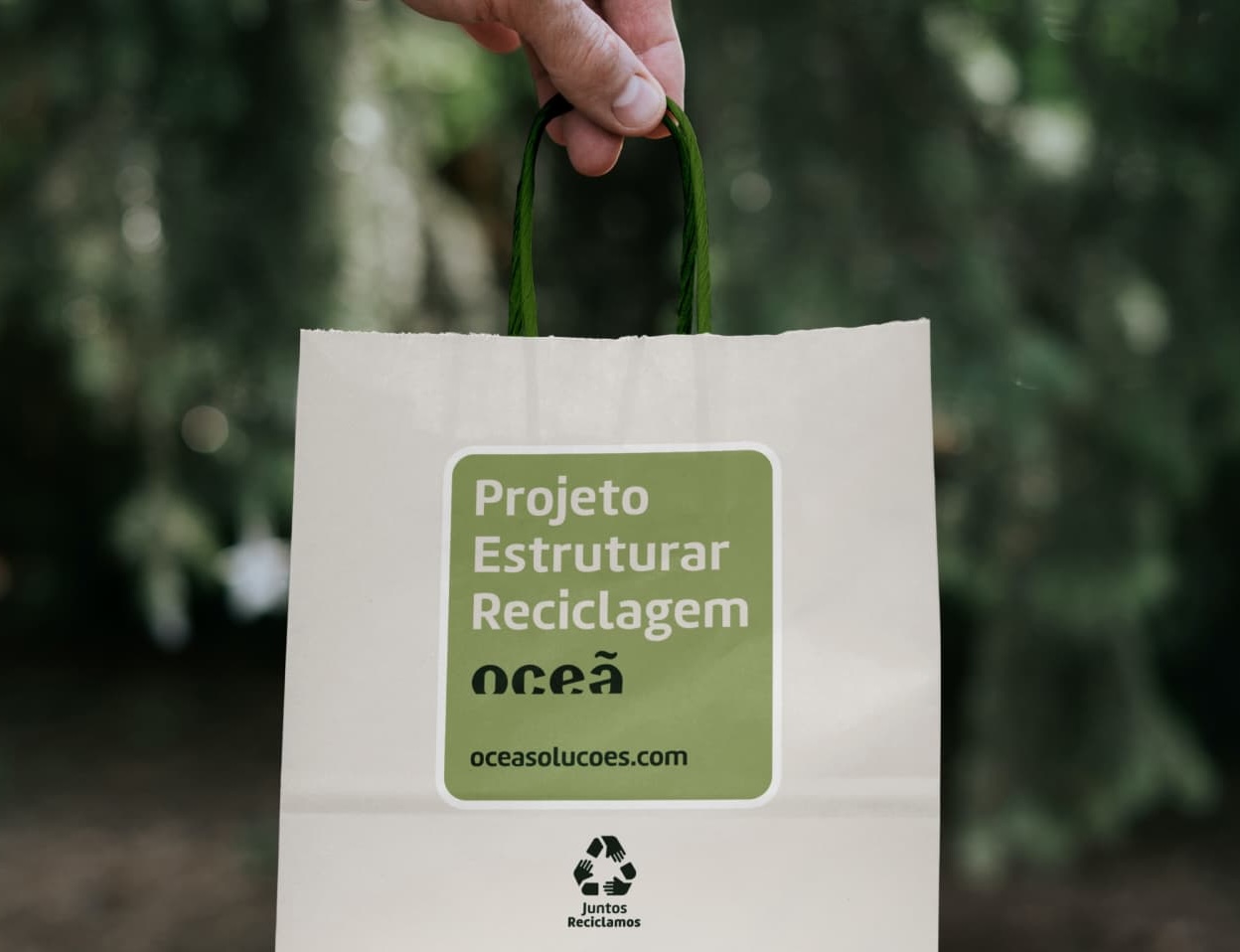 A imagem exibe uma mão segurando uma sacola de papel com a frase Projeto Estruturar Reciclagem Oceã, seguido do selo Juntos Reciclamos. Ao fundo vemos uma floresta de mata preservada.